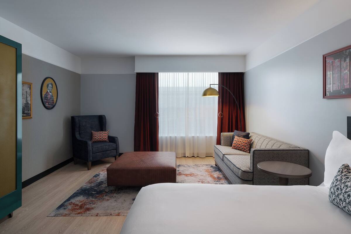Habitación de hotel con cama, sofá, sillón, mesita y ventana con cortinas.