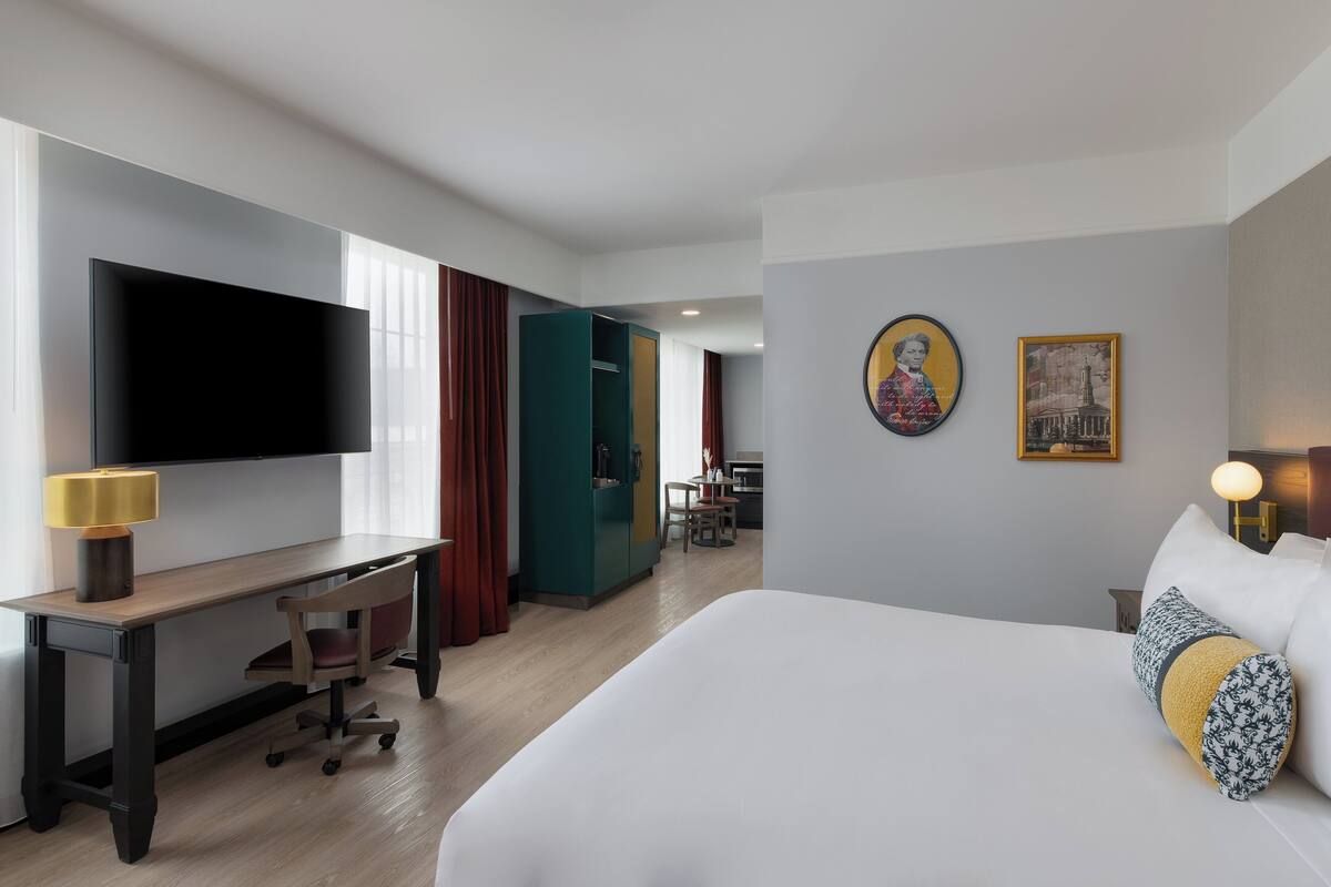 Habitación de hotel con cama, escritorio con silla, televisor y zona de comedor al fondo.