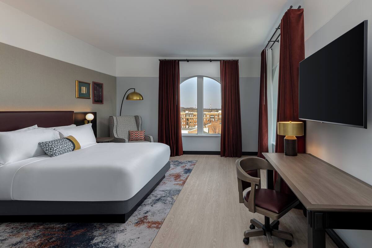 Habitación de hotel con una cama grande, un escritorio, una silla, un televisor y una ventana con cortinas.