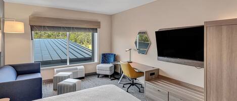 A hotel room with a bed, a desk, a chair, a TV, and a window with a view of the roof.