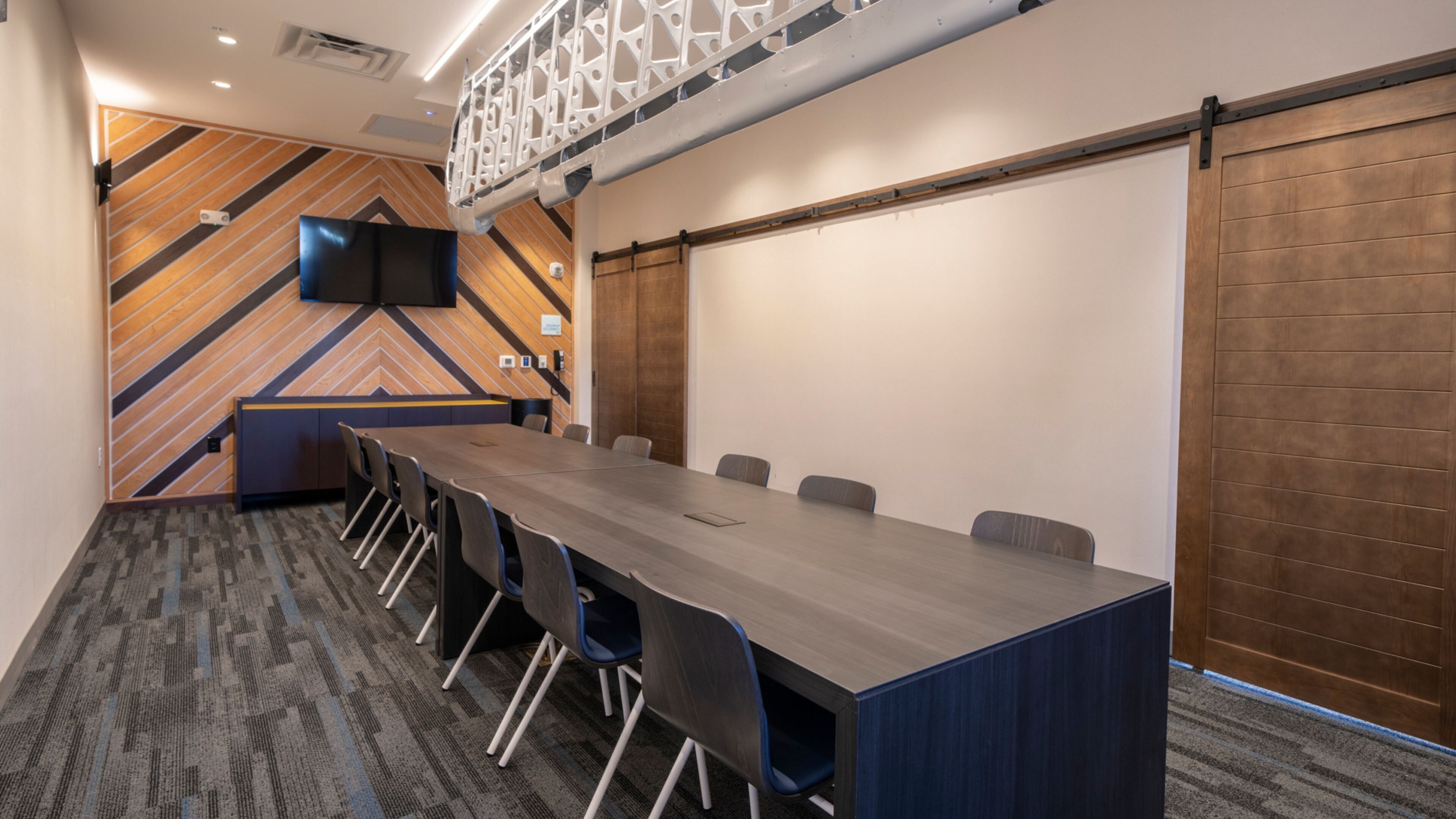 Ein Konferenzraum mit langem Tisch, Stühlen, einem Fernsehbildschirm und einer Holzwand mit geometrischem Muster.