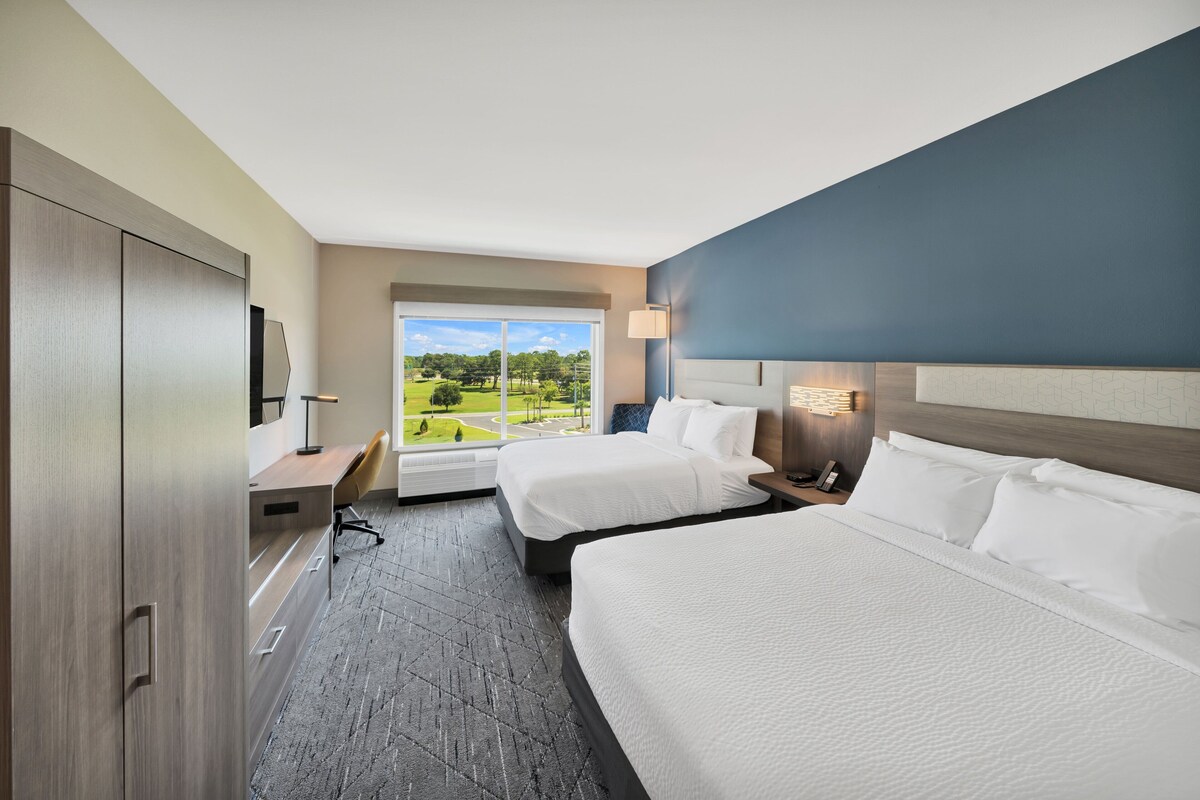 Une chambre d’hôtel avec deux lits, un bureau, une télévision et une fenêtre donnant sur un paysage verdoyant.