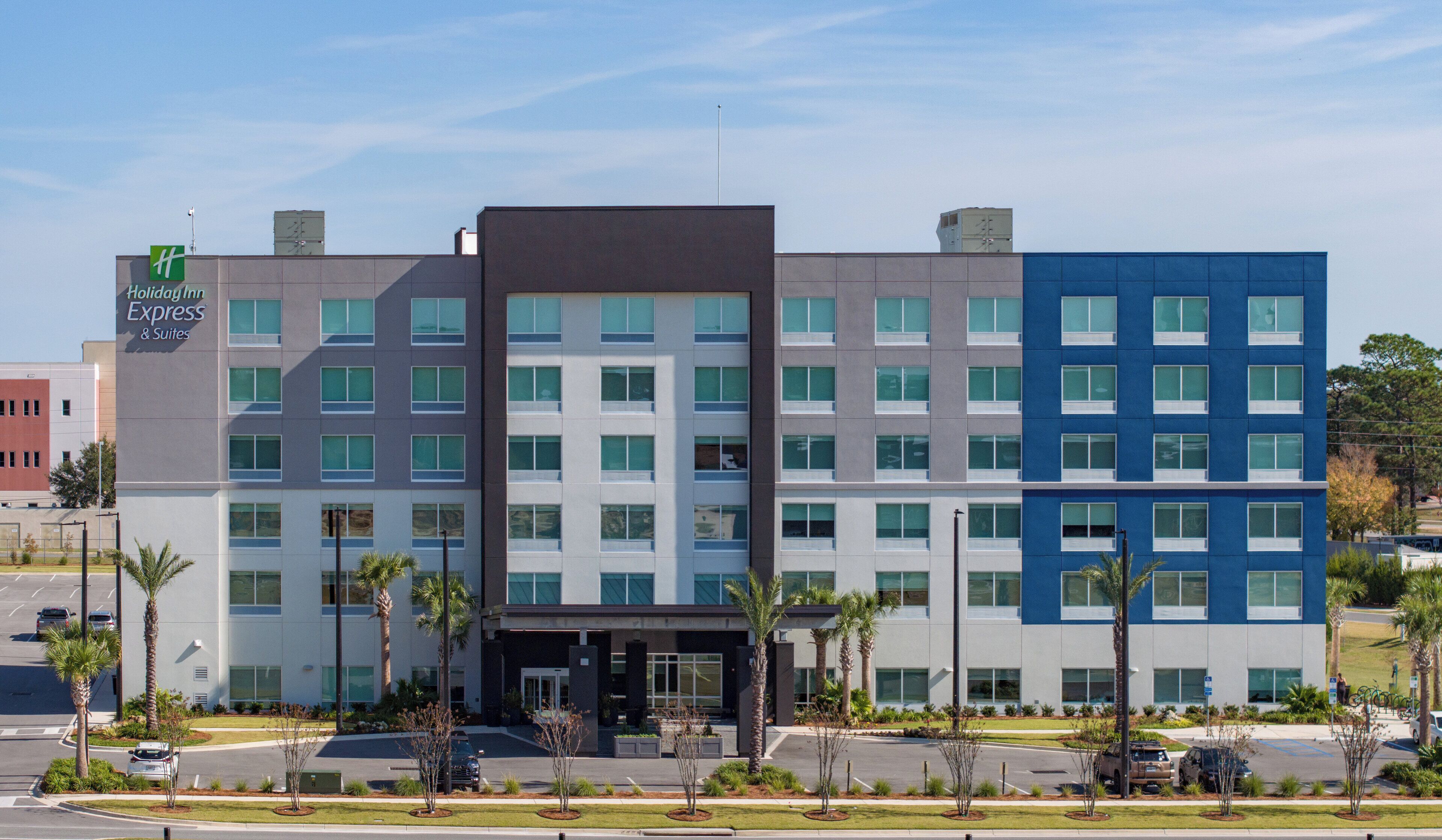 Edificio de Holiday Inn Express con diseño moderno, que presenta una combinación de fachadas grises y azules, numerosas ventanas y un estacionamiento con palmeras.