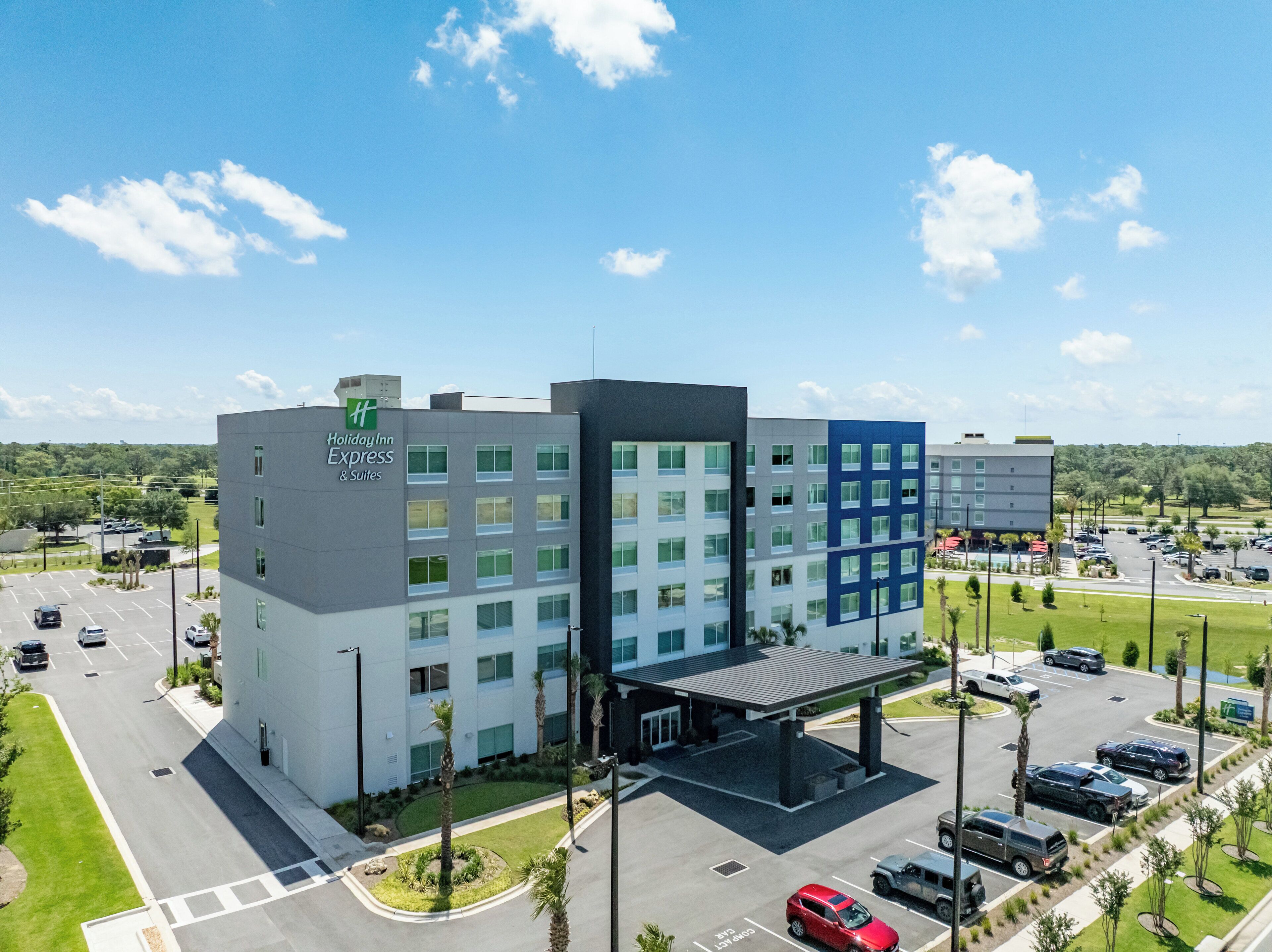 Un bâtiment Holiday Inn Express & Suites au design moderne, comprenant plusieurs étages, un parking couvert et un jardin aménagé à l’avant.