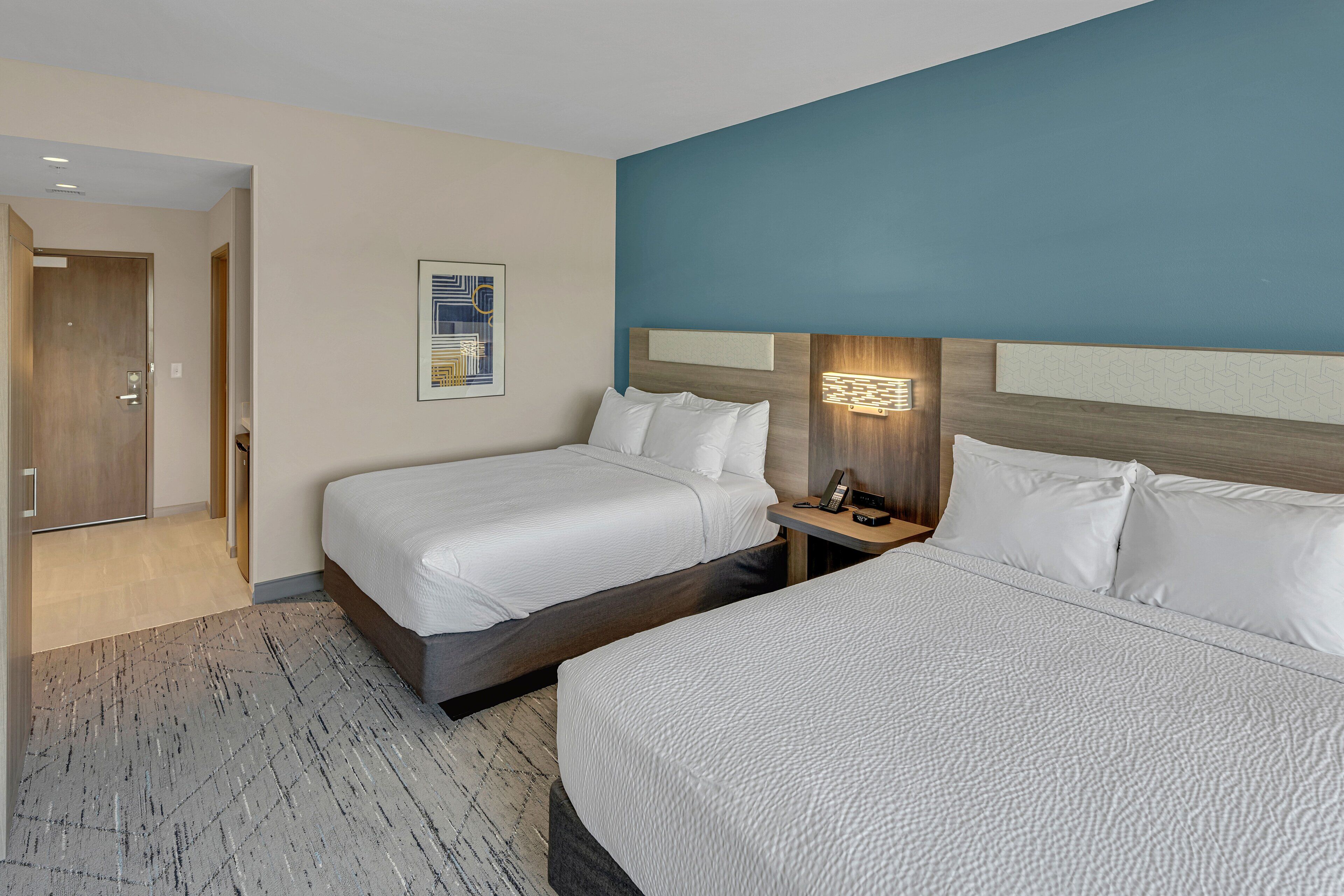 A hotel room with two beds, a nightstand, a wall-mounted lamp, and a door leading to another room.