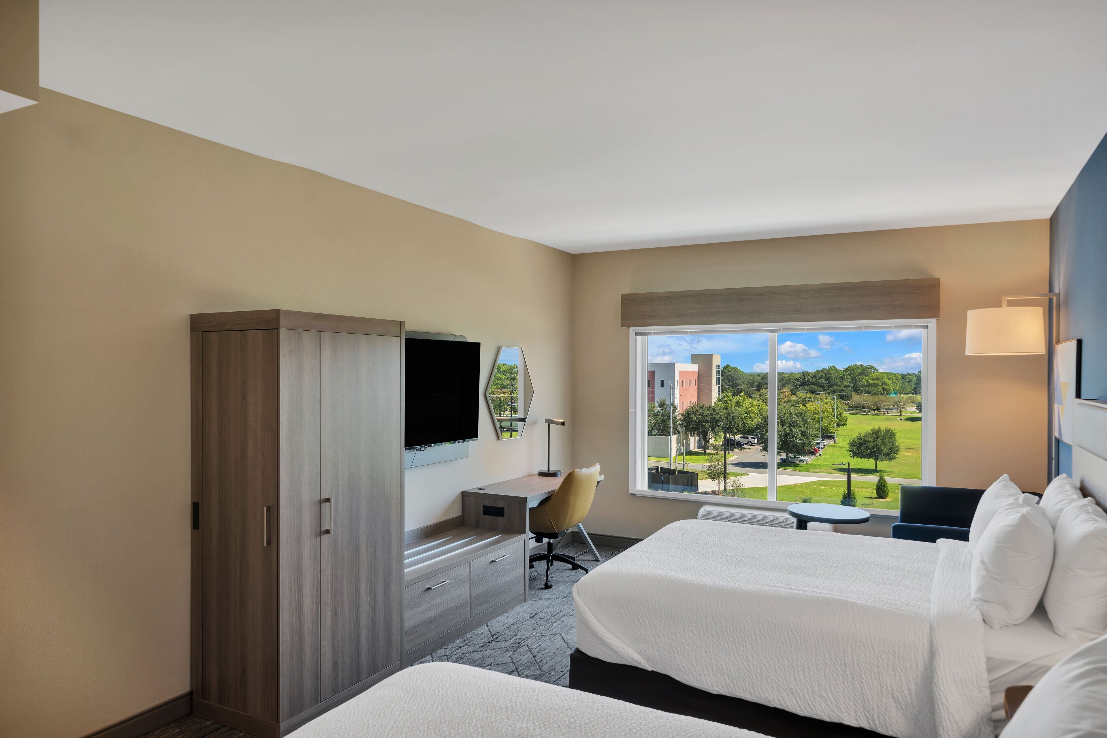 Ein Hotelzimmer mit zwei Betten, einem Schreibtisch, einem Stuhl, einem Fernseher und einem großen Fenster mit Blick auf einen Golfplatz und Gebäude.
