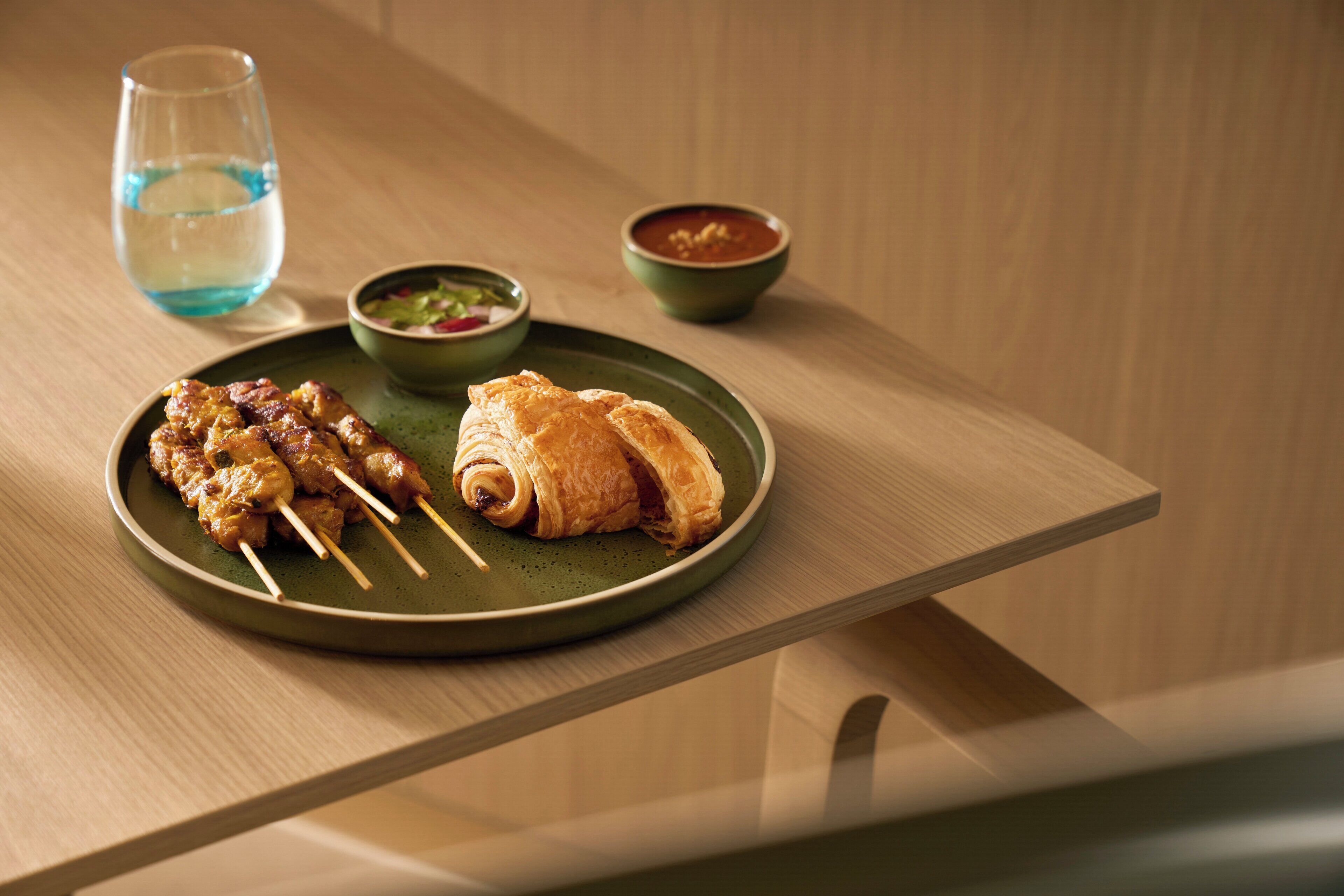 A plate with skewers and a croissant on a green plate, accompanied by two bowls of soup and a glass of water on a wooden table.