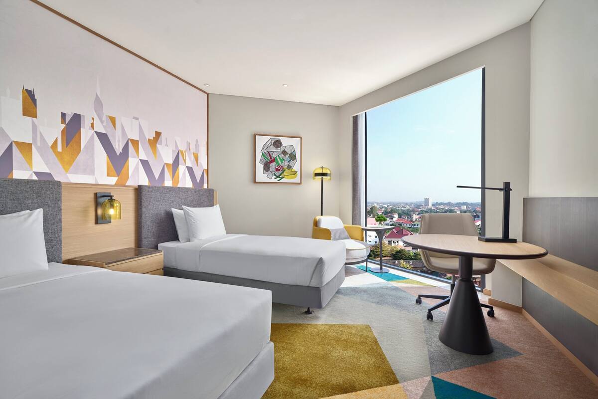 A modern hotel room with two beds, a desk, and a large window offering a city view.