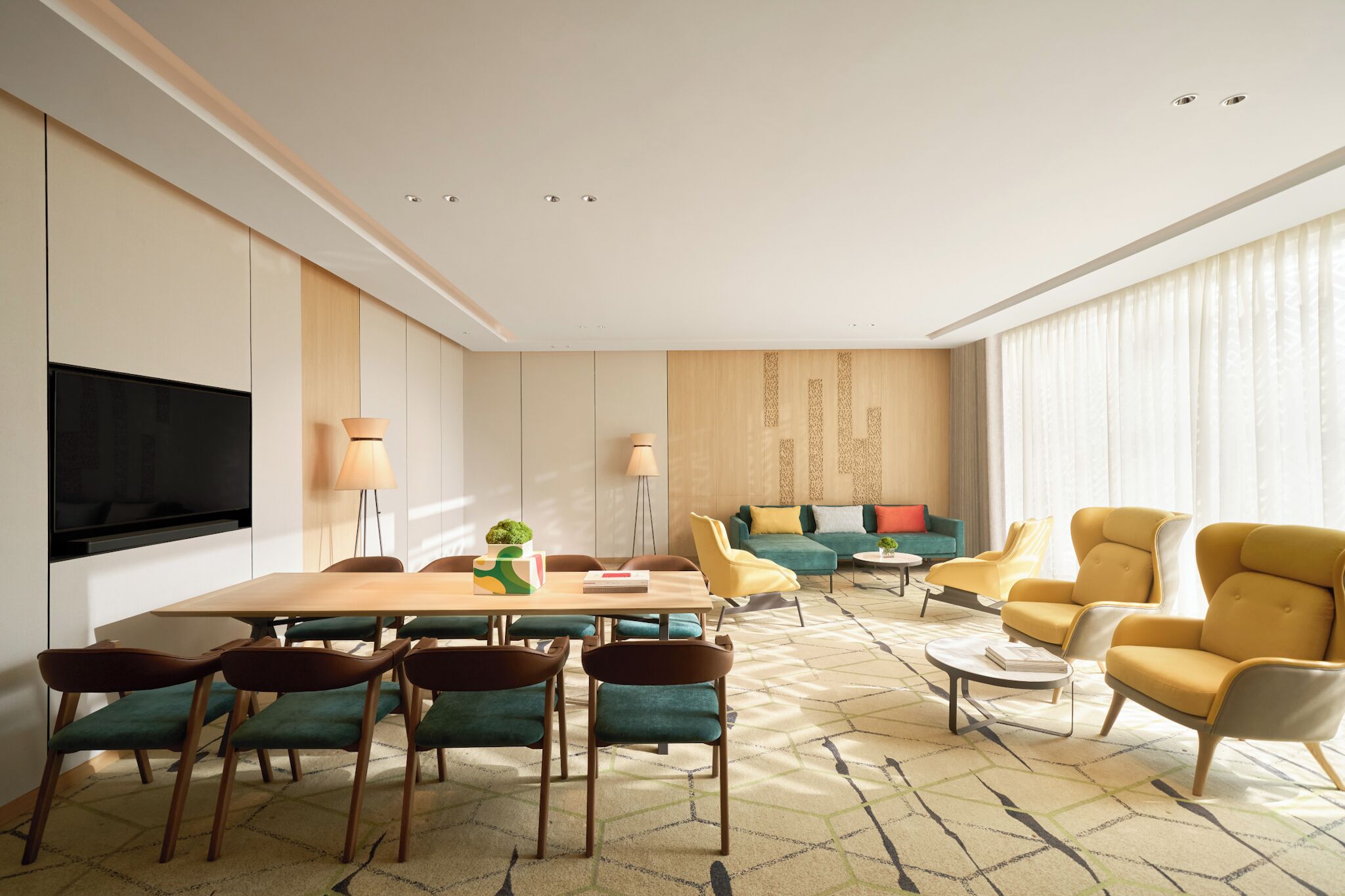 Una sala moderna con una mesa de comedor de madera, sillas tapizadas de color verde, un sofá con cojines amarillos y turquesas, y un televisor de pantalla plana montado en la pared.