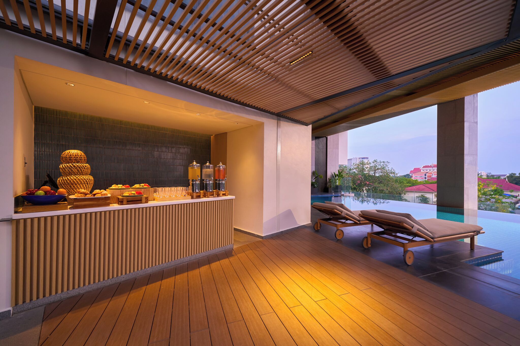A modern outdoor area with a wooden reception desk, a fruit bowl, and a poolside lounge with sun loungers.