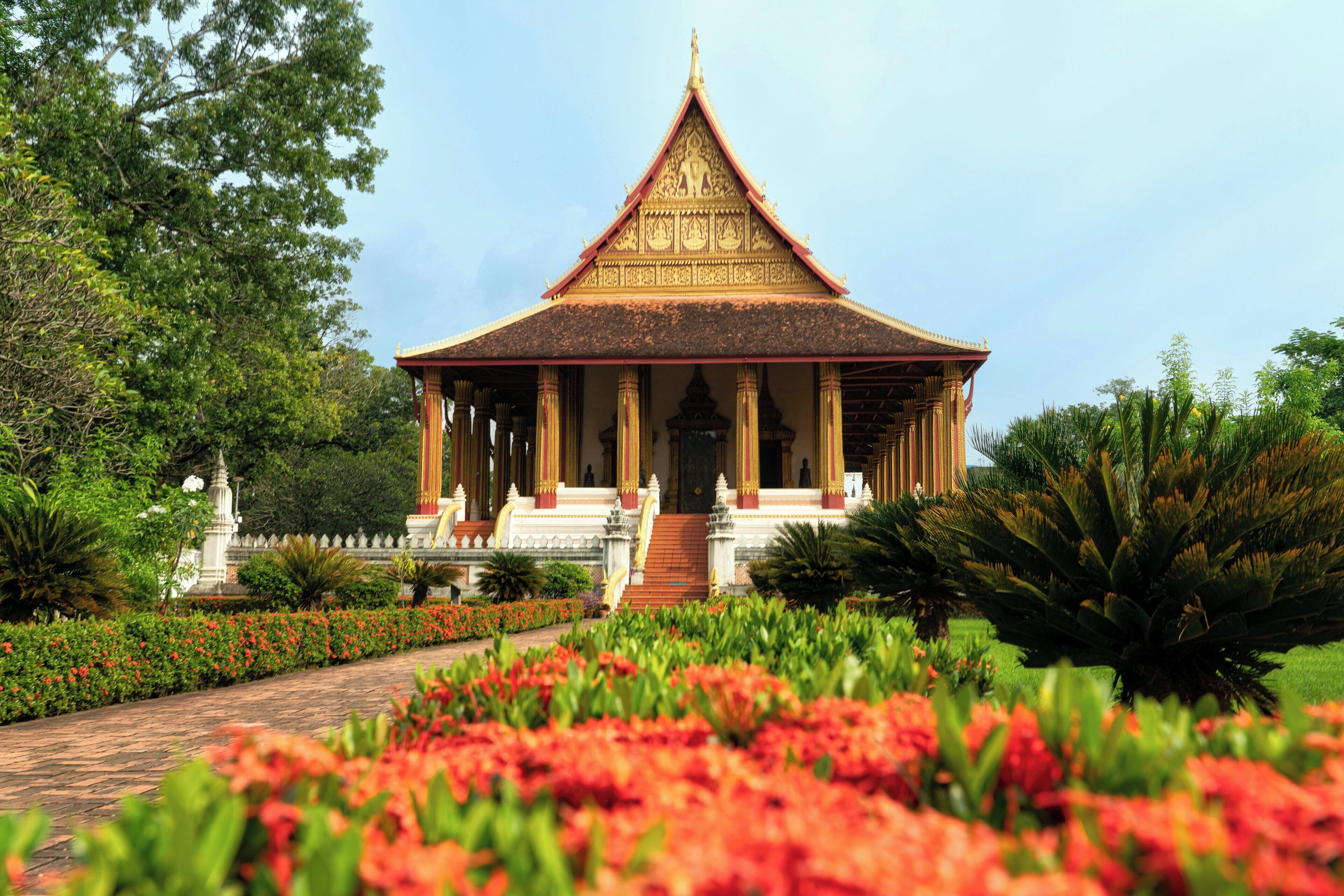 Een traditionele Thaise tempel met een terrasvormig dak, omgeven door weelderige begroeiing en kleurrijke bloemen.