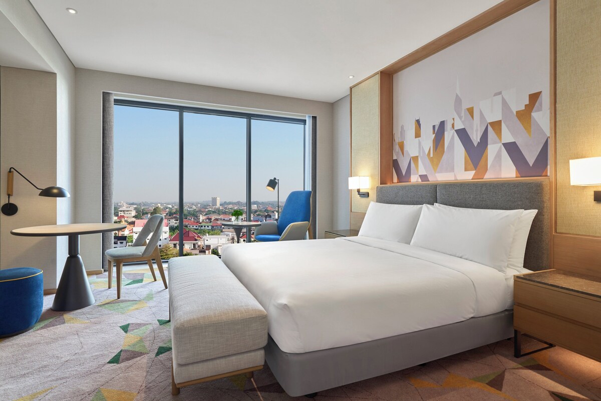 Habitación de hotel moderna con una cama grande, un escritorio y una silla. Se ve la ciudad a través de amplios ventanales.
