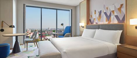 A modern hotel room with a large bed, a desk, and a chair. There is a view of the city through large windows.