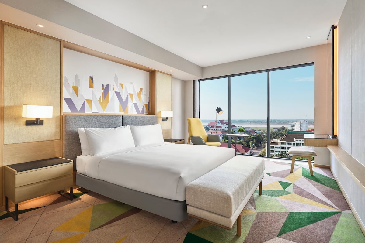 A modern hotel room with a large bed, two bedside tables, a chair, a desk, and a large window offering a city view.