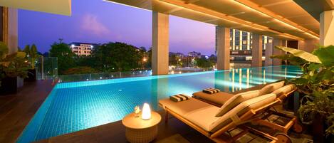 Piscina infinita moderna con sillones de descanso y una pequeña mesa, rodeada de plantas en macetas.