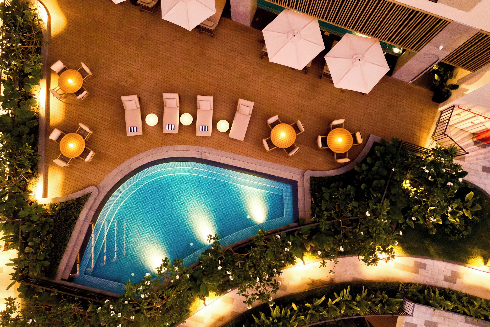 An aerial view of a rooftop pool area with sun loungers, umbrellas, and a swimming pool surrounded by greenery.