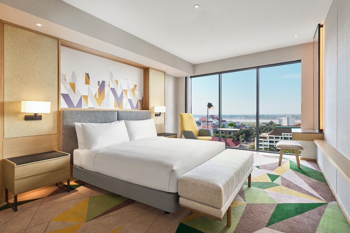 Una habitación de hotel moderna con una cama grande, dos mesitas de noche, una silla, un escritorio y un ventanal con vistas a la ciudad.