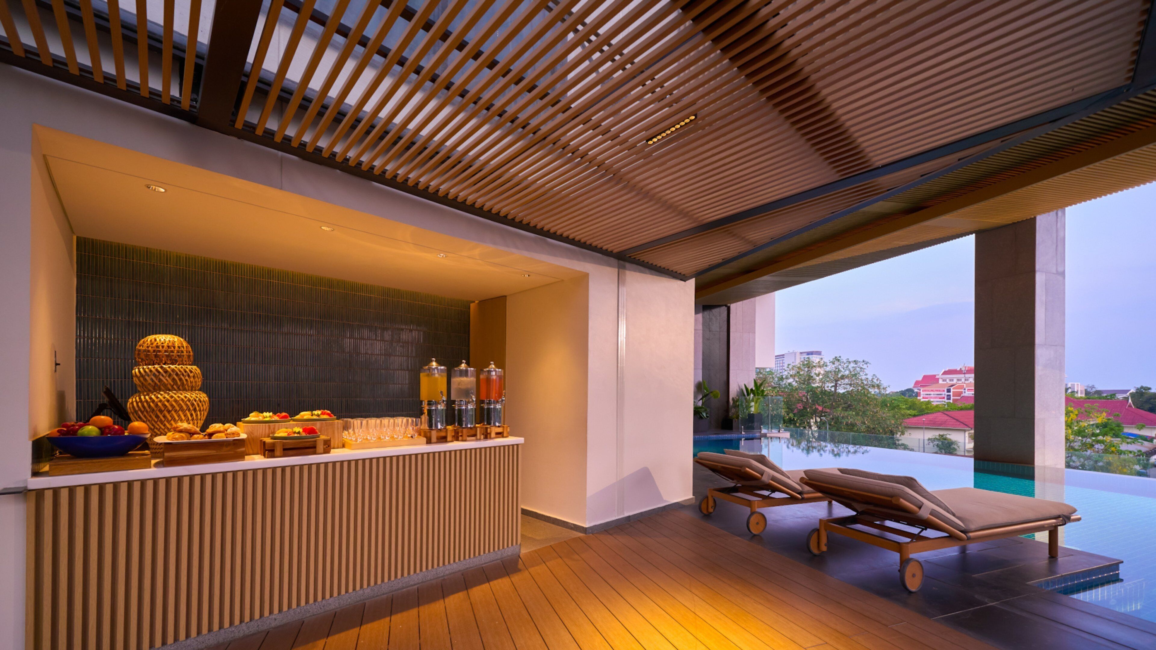 A modern bar area with a wooden ceiling, a fruit bowl, and a glass display. There are lounge chairs by a pool with a view of buildings and trees.