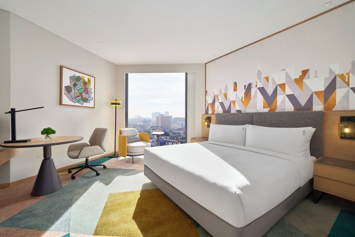 A modern hotel room with a large bed, a desk, a chair, and a view of the city.