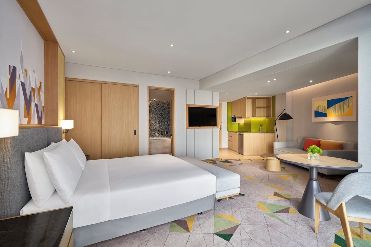 A modern hotel room with a large bed, a dining area, and a kitchenette.