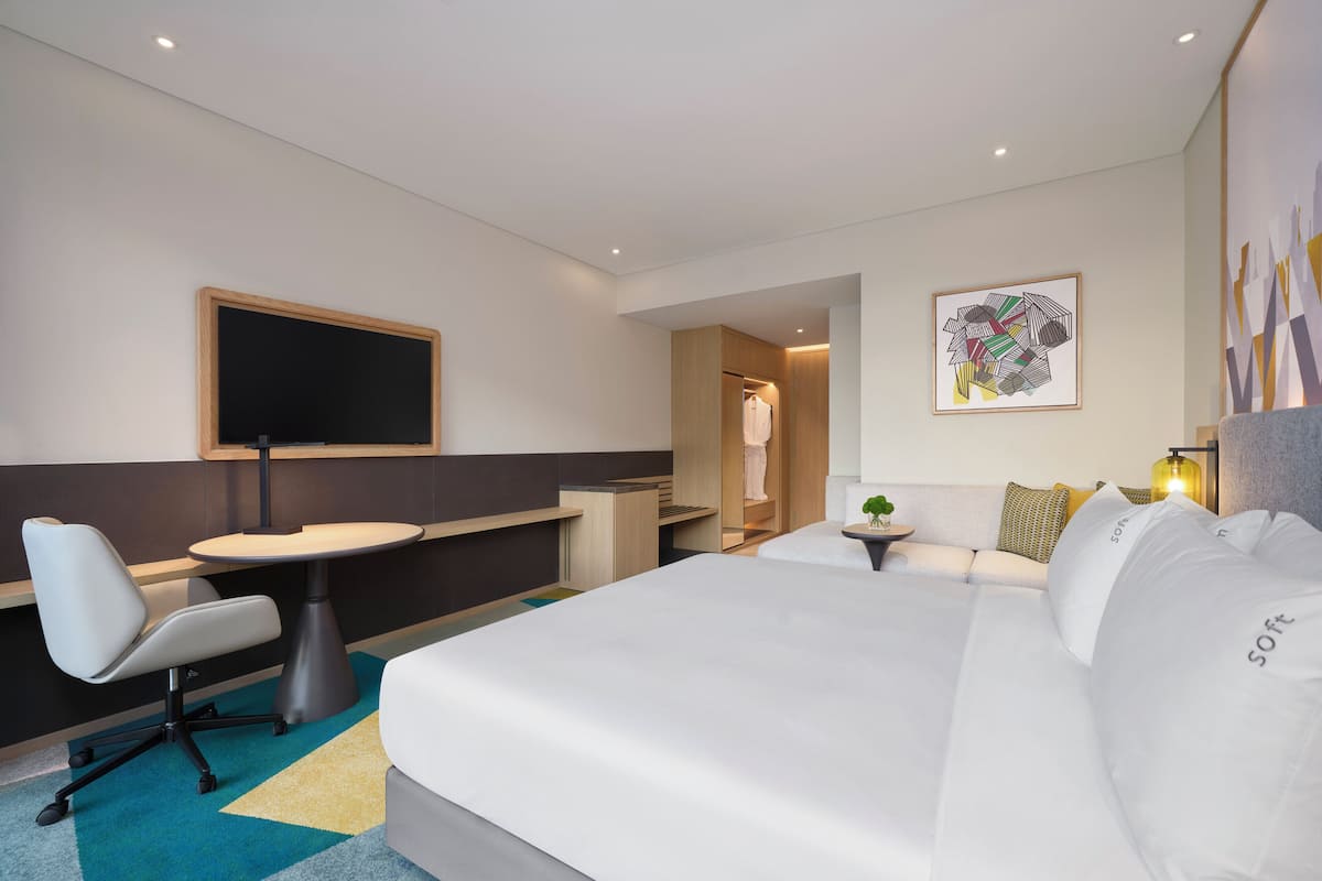A modern hotel room with a bed, a desk, a chair, a small table, and a bathroom visible through an open door.