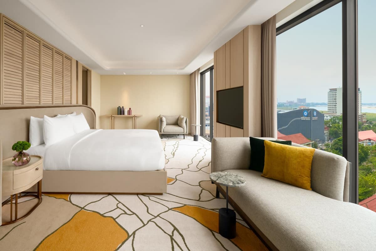 A modern hotel room with a large bed, a sofa, a TV, and a view of the city.