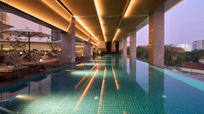 Una piscina interior moderna con un diseño largo y estrecho, rodeada de sillones de descanso y sombrillas, y una vista panorámica de la ciudad al fondo.
