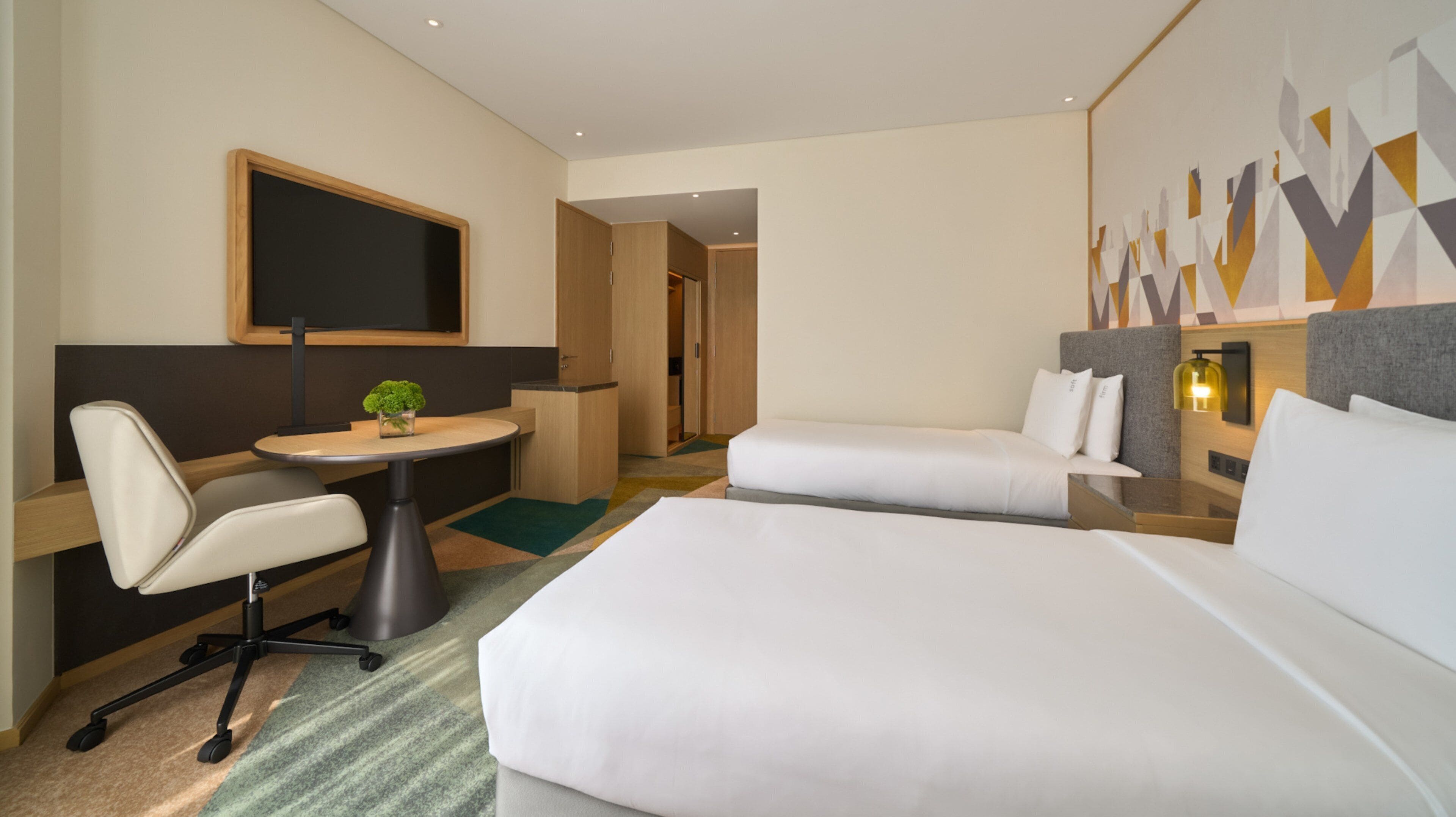 Une chambre d’hôtel équipée d’un lit, d’un bureau, d’une chaise, d’une petite table avec une plante et d’un téléviseur fixé au mur.