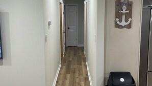 A narrow hallway with wooden flooring, white walls, and a door at the end. A black trash bin is placed against the wall on the right. - Gorgeously remodeled Seaside Oasis in Atlantic Beach! (Atlantic Beach)