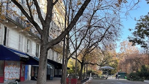 Una calle arbolada con un banco, un edificio con toldo azul y una zona de juegos.