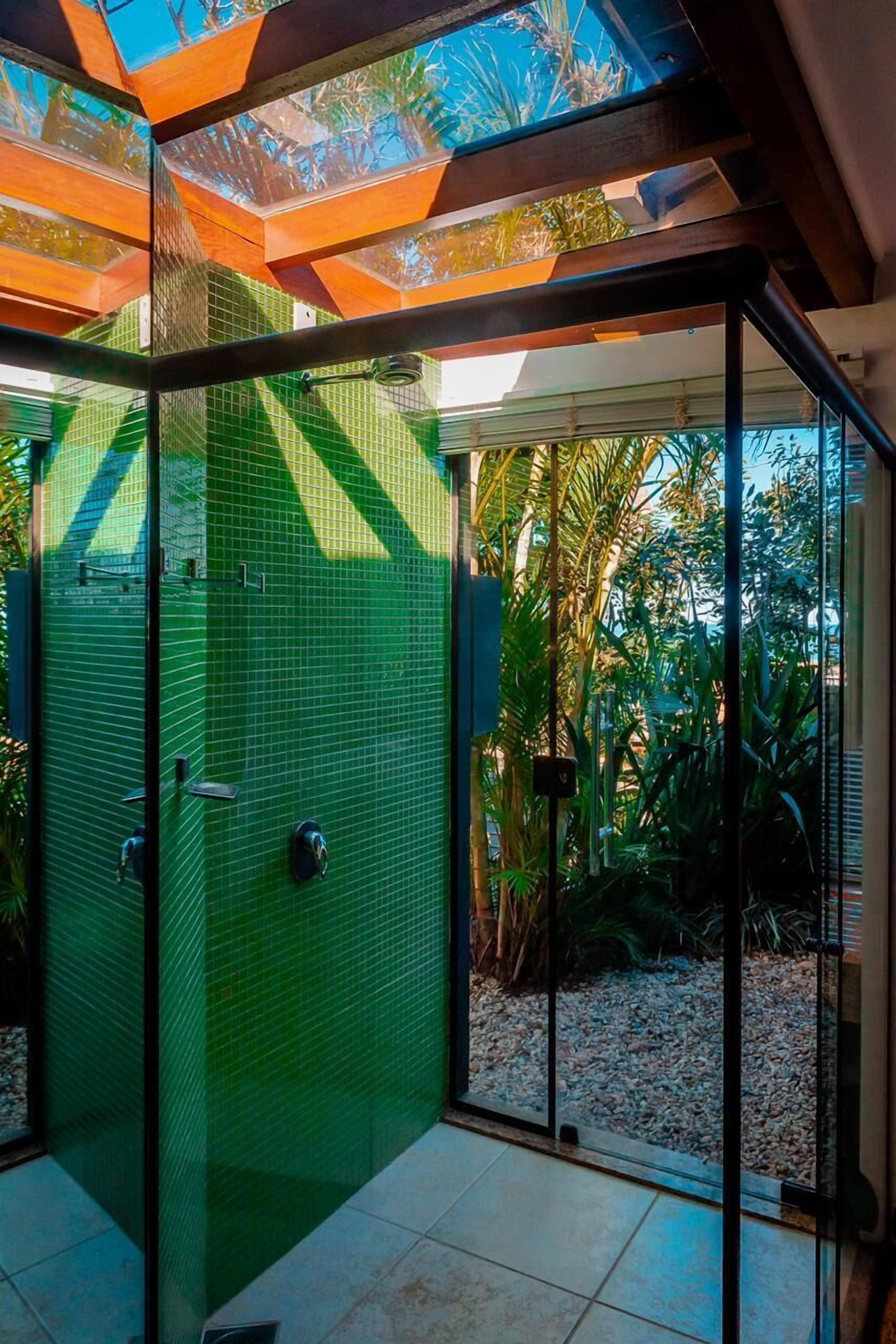 A modern glass-enclosed shower area with a green tiled wall and a view of tropical plants.