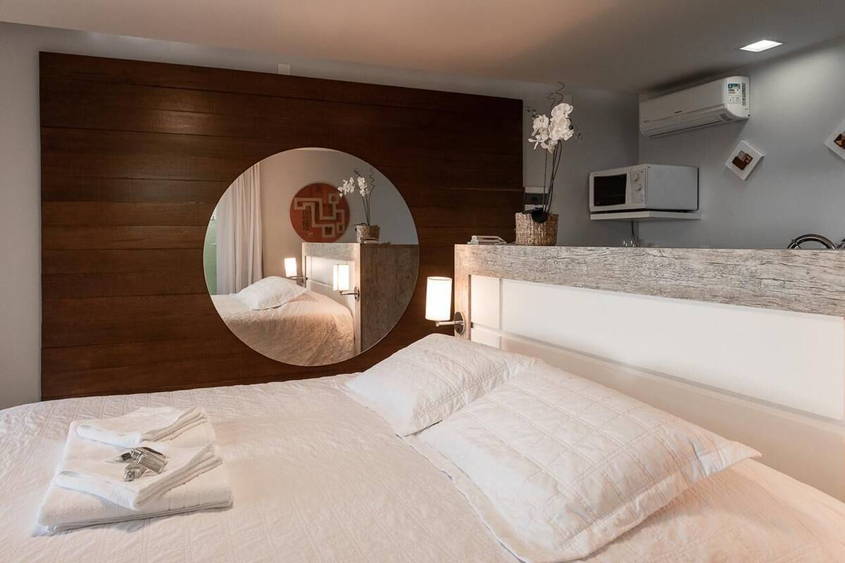A modern hotel room with a large round mirror, a wooden headboard, a white bed with neatly folded towels, a built-in TV, and a small side table with a vase of flowers.
