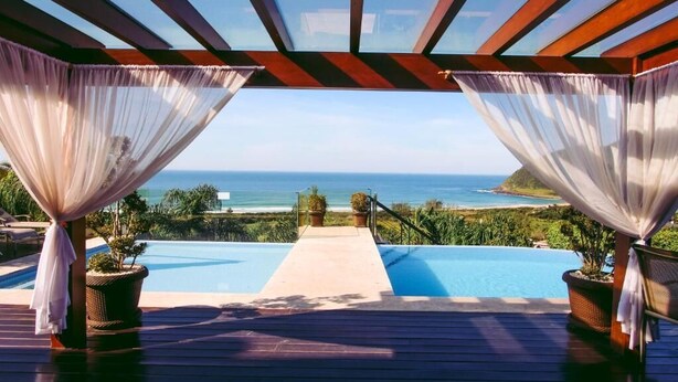An infinity pool with a view of the ocean, surrounded by a wooden deck and draped with sheer curtains. - Morada Prainha (Garopaba)