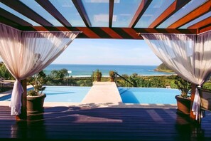 Una piscina infinita con vista al océano, rodeada por una terraza de madera y con cortinas de gasa.