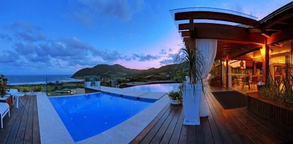 Zona de piscina moderna con vista al océano y las colinas al anochecer.