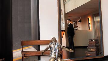 A room with a wooden bench, a small table with magazines, and a large mirror reflecting a staircase and a person.
