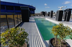 Un espace piscine aménagé sur le toit, avec un bâtiment bleu, des chaises longues et des plantes en pot.