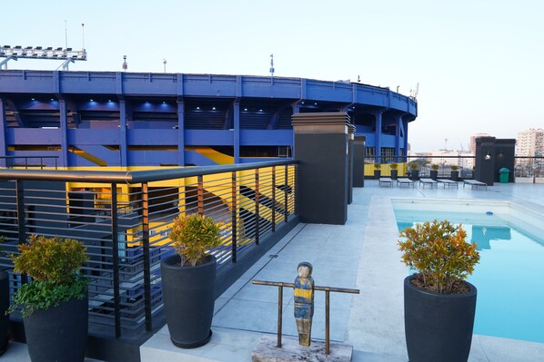 Un espace piscine aménagé sur le toit, avec un bâtiment aux couleurs bleue et jaune, des plantes en pot et une statue.