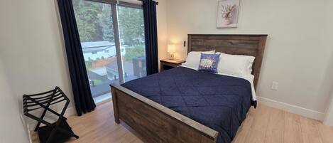 Ein Schlafzimmer mit einem hölzernen Bett, einem Nachttisch, einer Lampe, einem Fenster mit Vorhängen und Blick ins Freie.