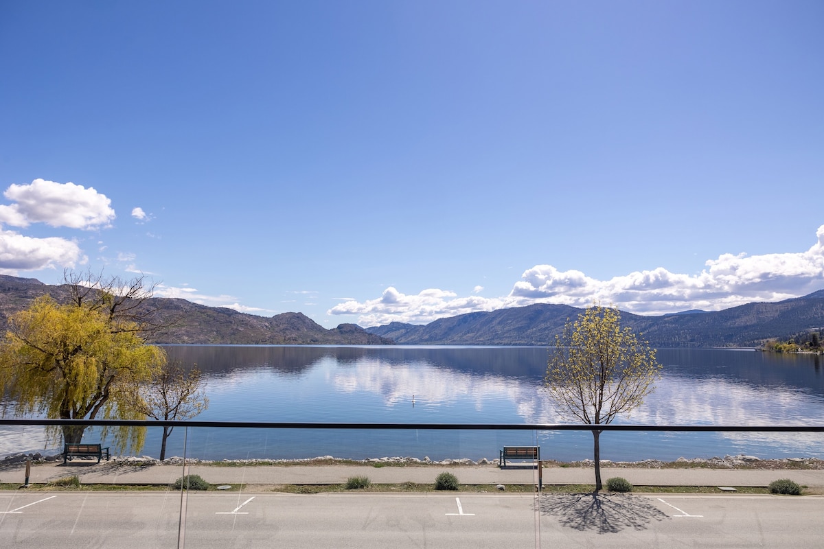 Um estacionamento com um banco e uma árvore em frente a um lago calmo, com montanhas ao fundo.