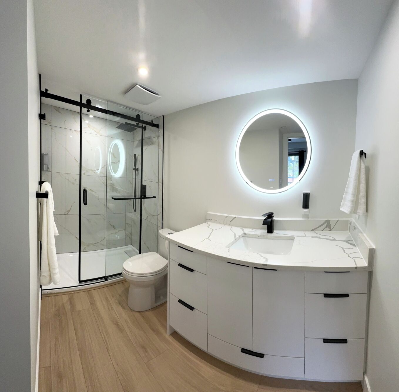 Un baño moderno con cabina de ducha de vidrio, tocador blanco con espejo circular y un inodoro.