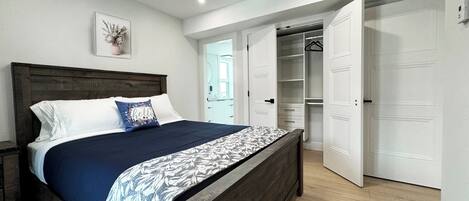 A bedroom with a wooden bed, white walls, a ceiling light, and an open closet with white doors.
