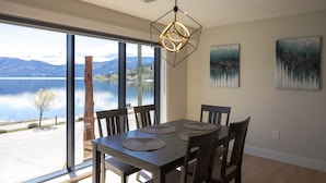 Uma área de refeições com mesa posta para quatro pessoas, janelas amplas com vista para um lago e montanhas, e duas pinturas abstratas na parede.