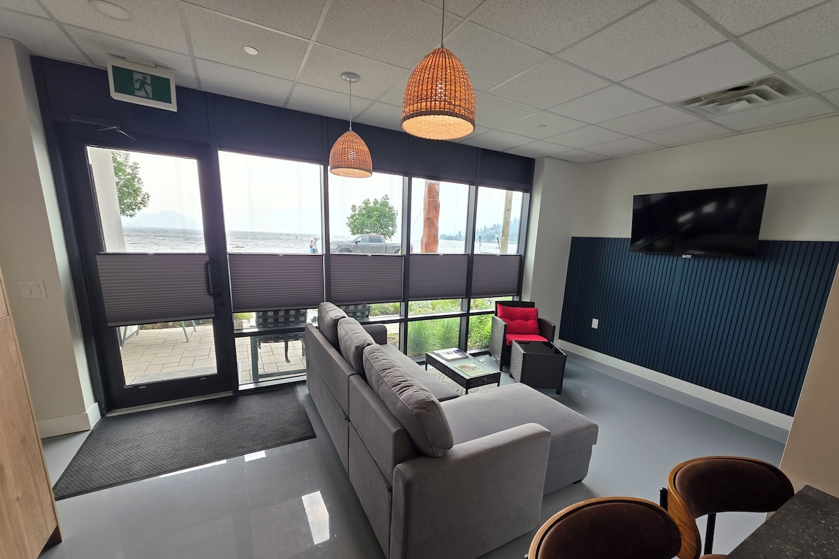 A modern office space with a sofa, chairs, a TV, and large windows overlooking a body of water.