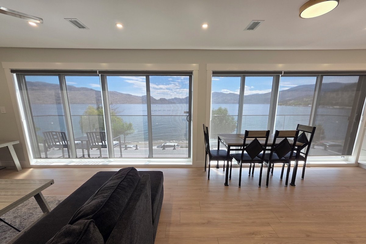 Quarto espaçoso com uma grande porta de vidro deslizante que dá para uma varanda com vista para um lago. No interior, há um sofá de cor escura e uma mesa de jantar com cadeiras.