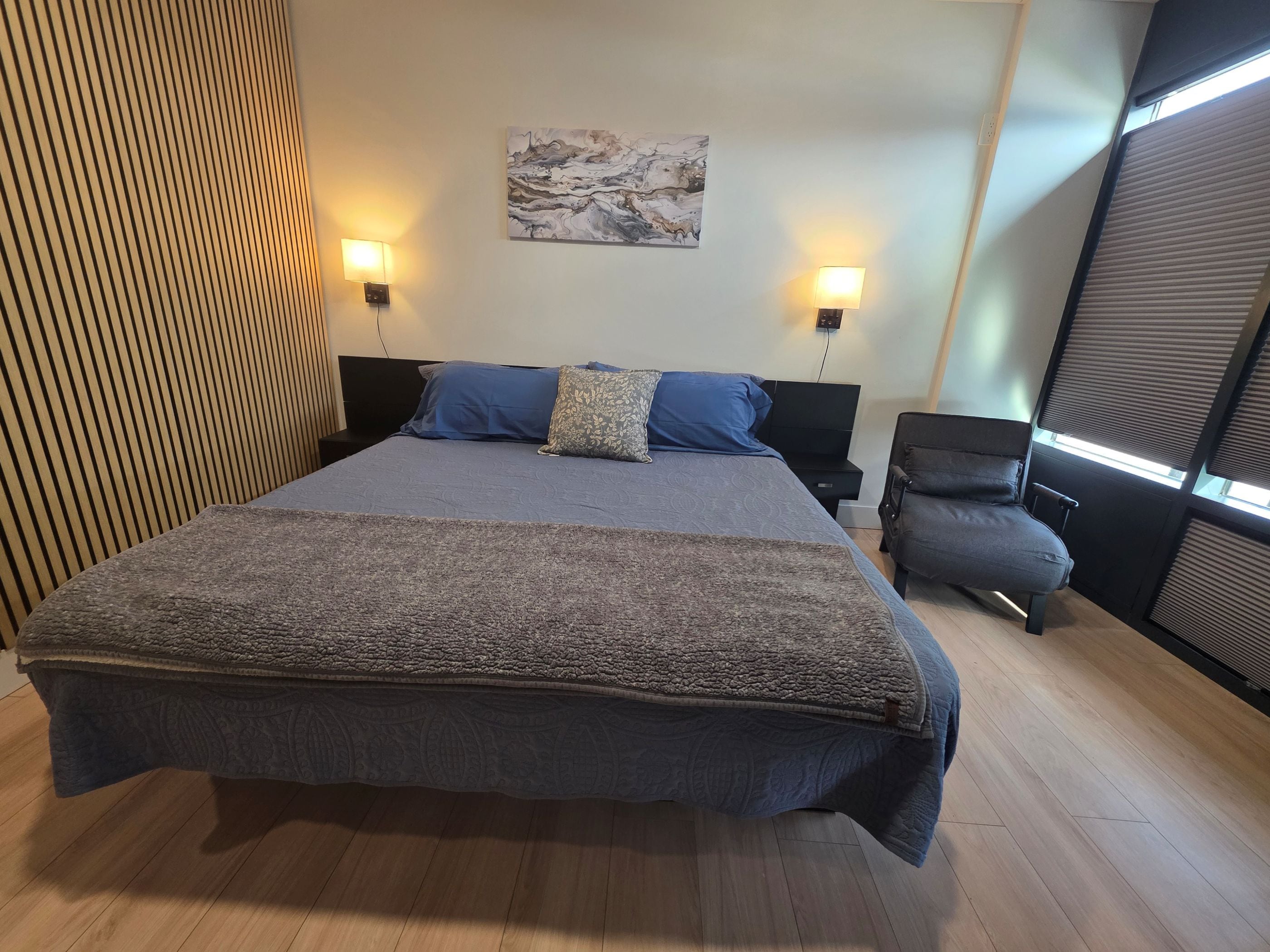 A large bed with a patterned bedspread, two bedside lamps, a wall-mounted painting, and a window with blinds.