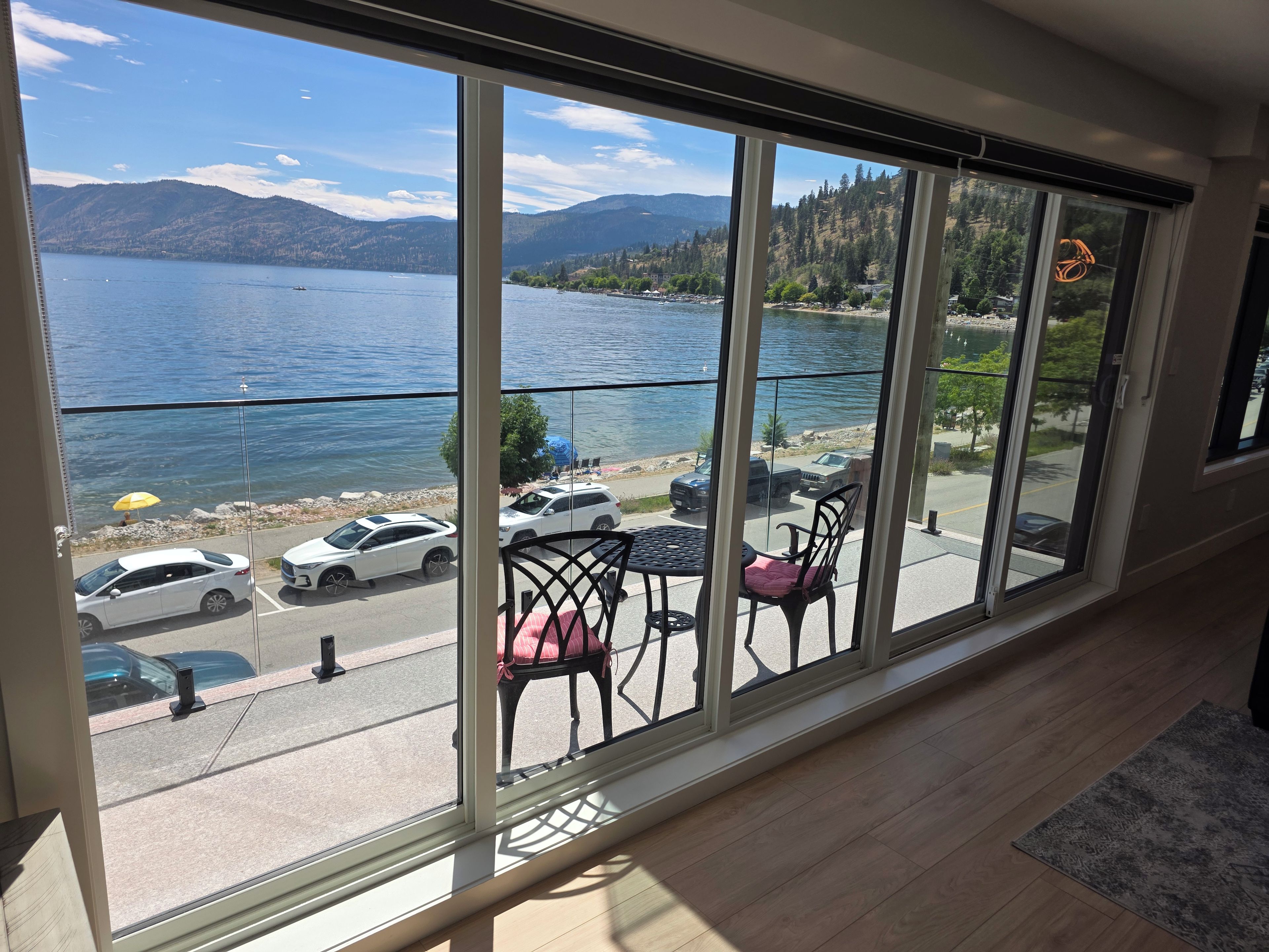 Habitación con una ventana grande que da a un lago y montañas, un balcón con sillas y una mesa, y vistas a coches aparcados y una playa.