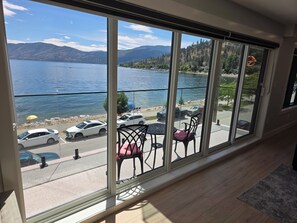 Una stanza con una grande finestra che si affaccia su un lago e montagne, un balcone con sedie e tavolo, e vista su auto parcheggiate e una spiaggia.