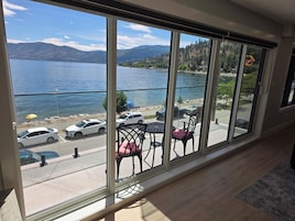 Habitación con una ventana grande que da a un lago y montañas, un balcón con sillas y una mesa, y vistas a coches aparcados y una playa.