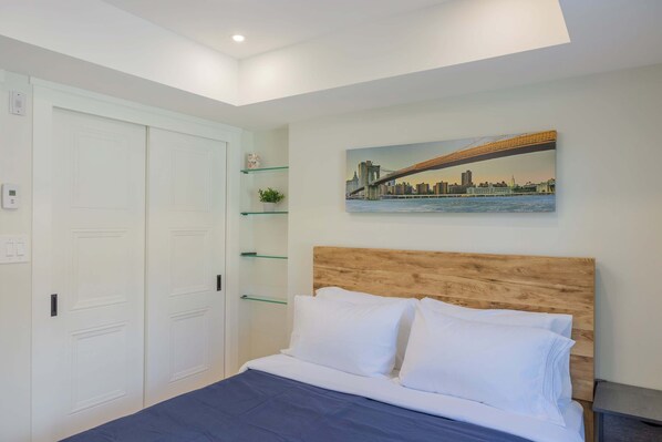 A bedroom with a bed, white walls, a wooden headboard, a painting of a bridge, and a closet with glass shelves.